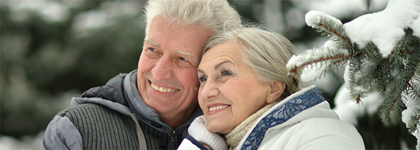 Elderly man and woman dressed in winter clothes smiling outside by a snowy tree