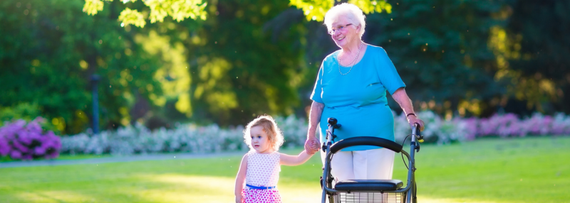 Happy senior lady with a walker rollator and a toddler girl walking together in the park in summertime
