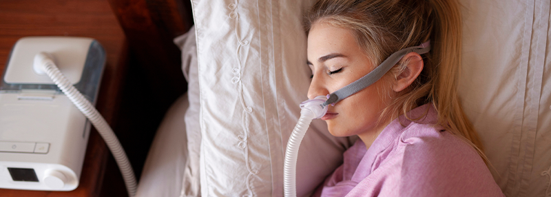 Woman sleeping on her side in bed with a CPAP mask on connected to a CPAP machine next to the bed
