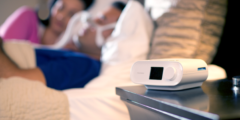Photo of a CPAP machine and a couple sleeping in the background.