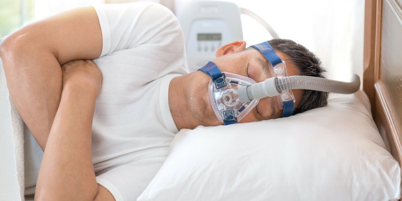 Photo of man in white shirt sleeping on a bed with a CPAP mask on his face.