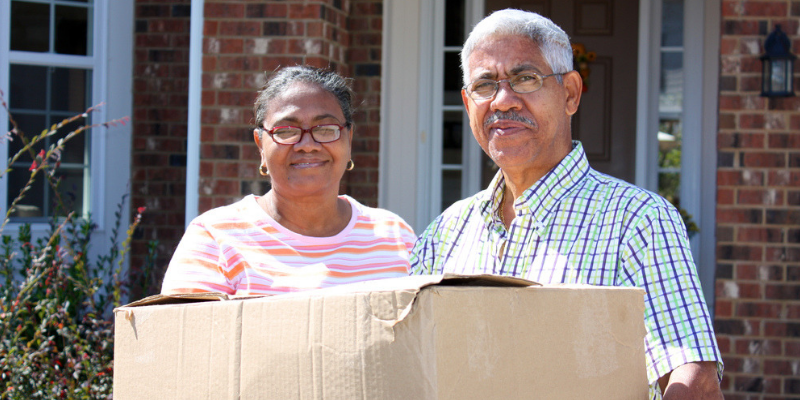 Photo of elderly couple standing outside a home with boxes in their hands.