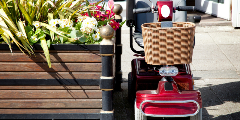 Picture of a red scooter with a basket on the front outside by some flowers and plants.