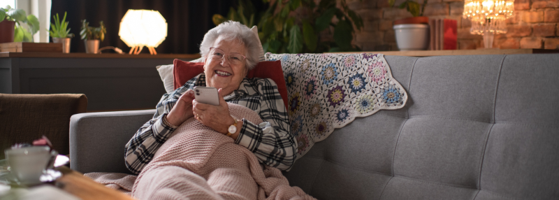 Senior woman sitting on couch under blanket smiling and using her phone