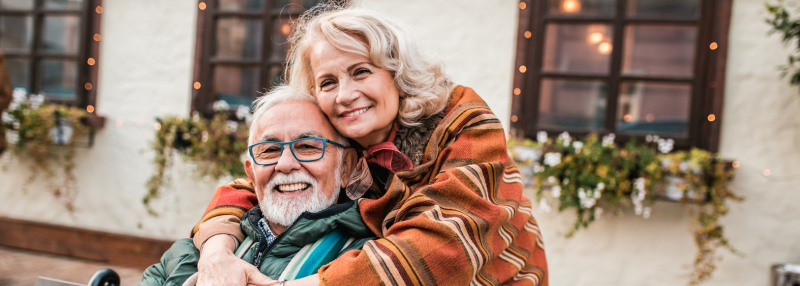 Senior man sitting on bench with his wife's hugging him from above