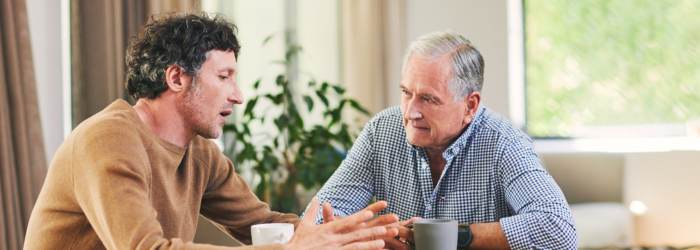 Adult son speaking with his senior dad at the dining table with coffee mugs